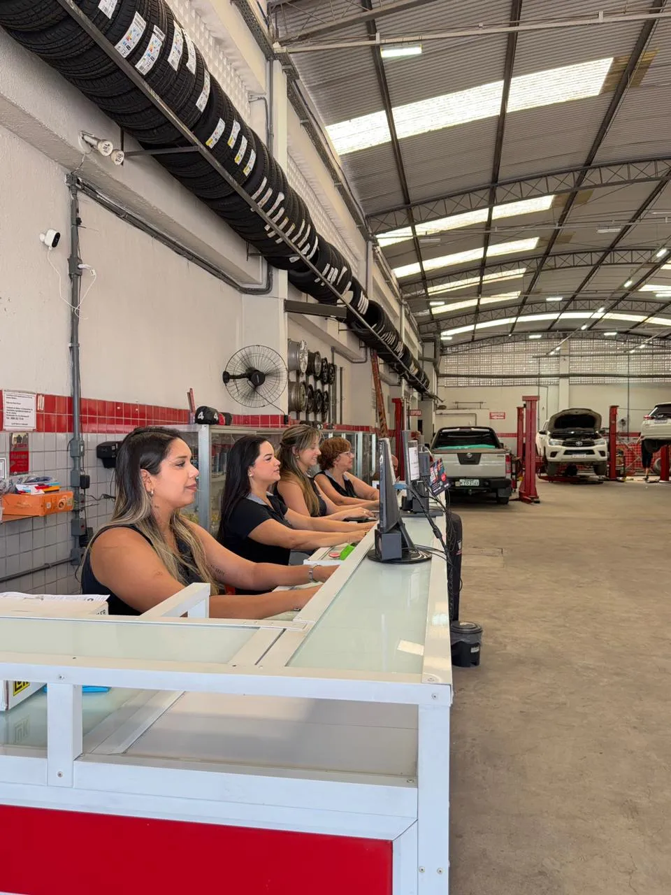 Equipe feminina de recepção da Magrão Auto Center no balcão de atendimento com oficina ao fundo em Campo Grande, RJ